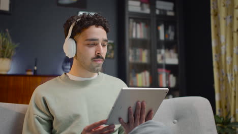 young man watching interesting movie on tablet while eating chips and sitting with crossed legs on couch at home