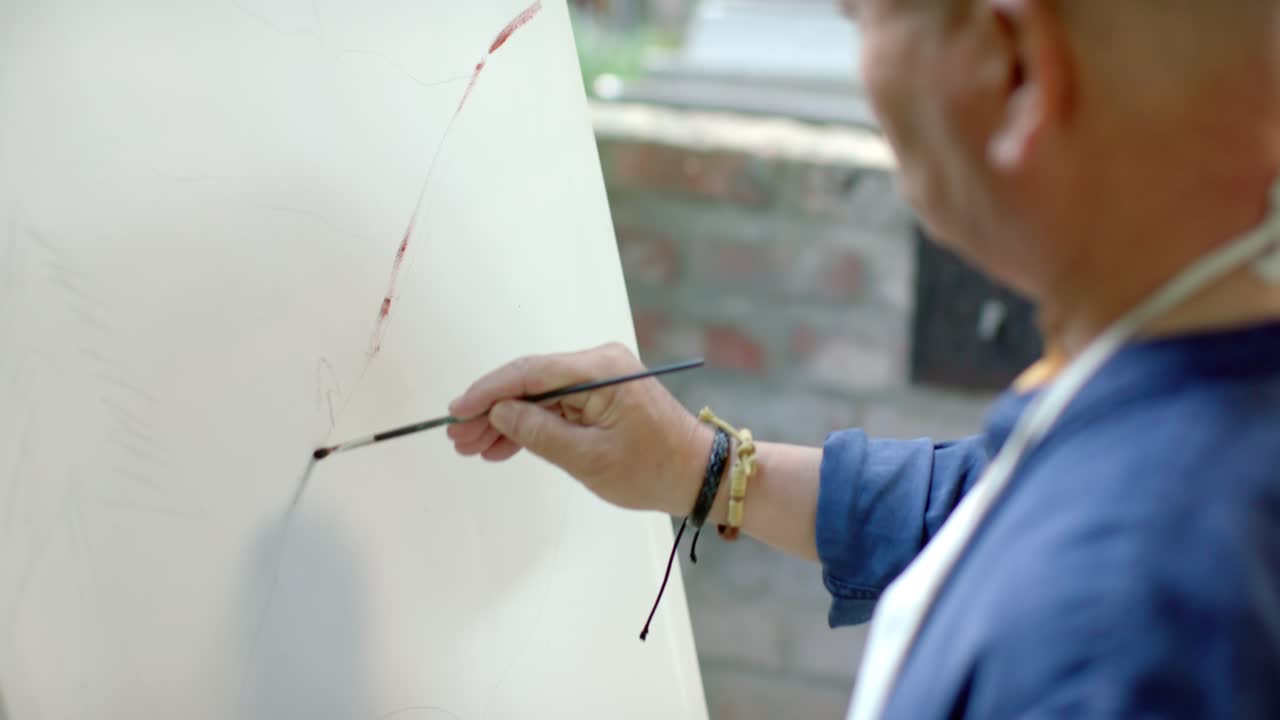 Hombre mayor pintando sobre lienzo en un patio, inspirado por una marca rojiza, mezclando la paleta, con espacio para texto.