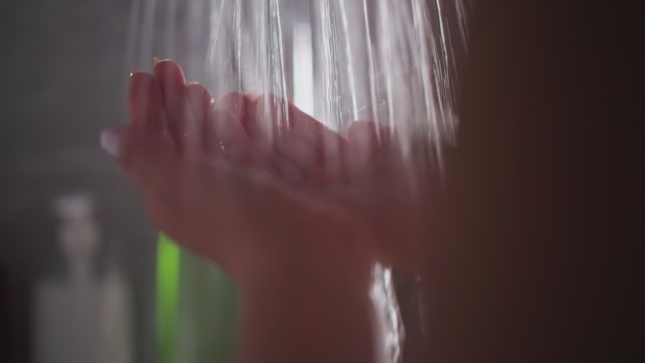 여자는 따뜻한 물의 폭포에서 위안을 찾습니다. 젊은 여자는 샤워에서 흐르는 하천 에서 손을 고 있습니다. 여자는 은 물 에 서 있습니다.