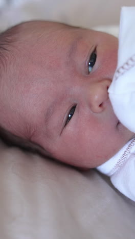 newborn kid in white costume lying on the bed and looking with his eyes wide-open. baby puts his hand in front of his mouth. close-up. vertical video