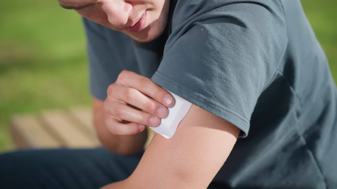 close up of white student seated outdoors pressing nicotine patch onto upper left arm while lifting shirt sleeve, with natural sunlight reflecting off skin and soft blur of green landscape in background