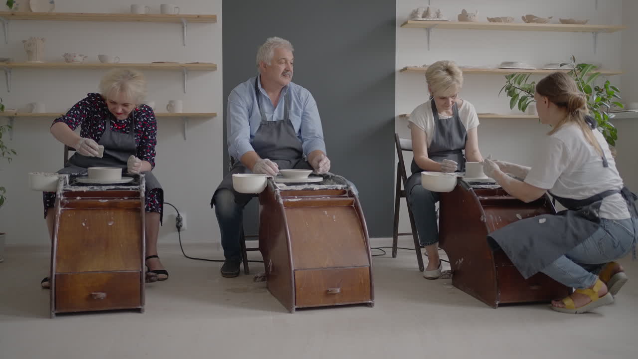 A female potter's wheel sculpting teacher explains how to work and teaches an elderly woman to work with clay and make mugs and jugs. Master class for pensioners. Pottery courses