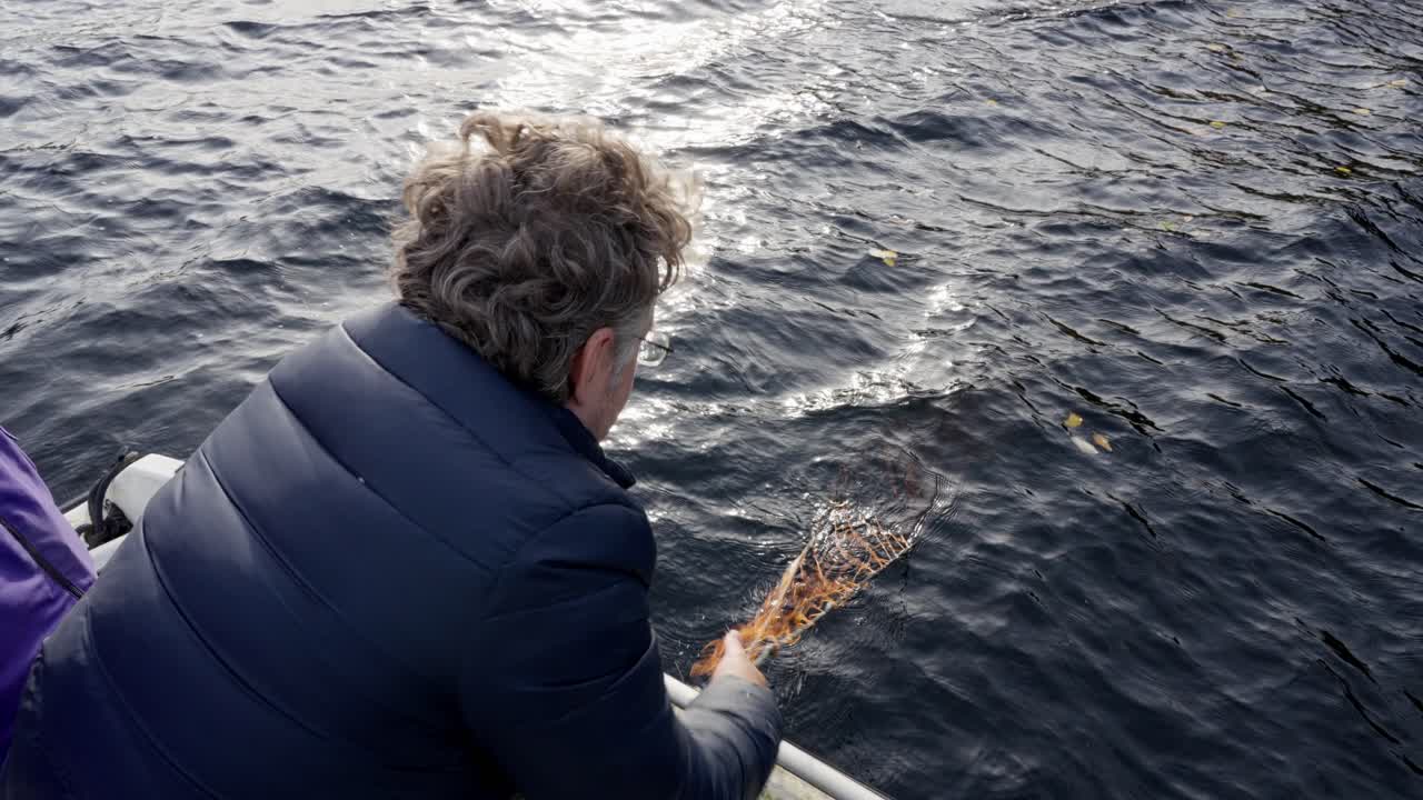 A man in his 40s hoists a crab net from the sea into a boat, handheld high angle