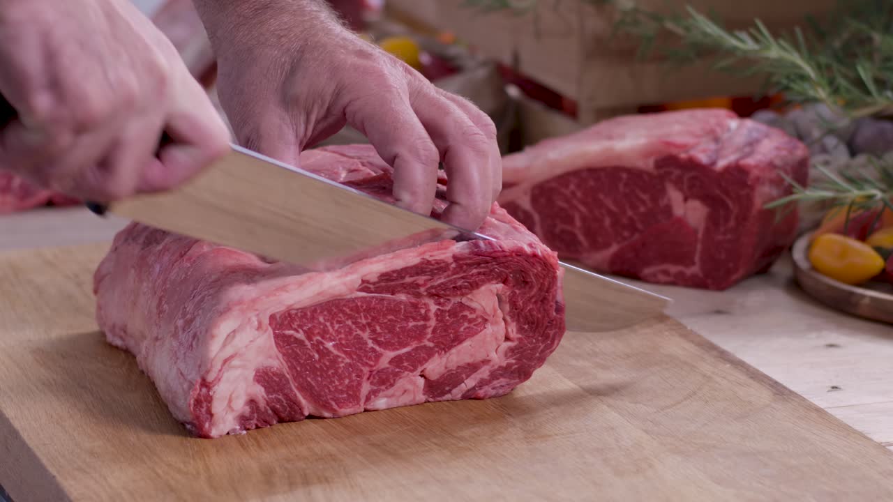 Close up of a butcher's hands using a sharp knife to carve a thick marbled ribeye steak from a prime beef roast on a wooden board