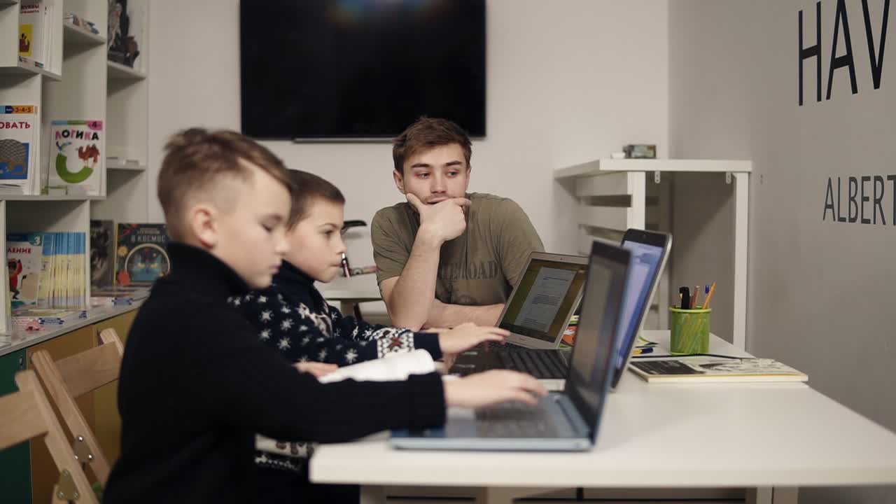 Young attractive male teacher in programming class helps two little boys learn something about gadgets such as laptops. Educational process.