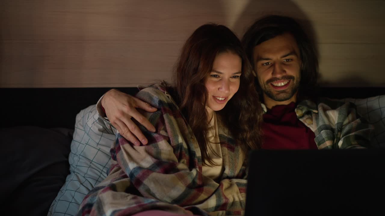 Happy brunette girl in a plaid shirt lies on the sofa in a trailer with her boyfriend with stubble and they watch a movie together using a laptop during their vacation in a camp outside the city in the summer with dim lights
