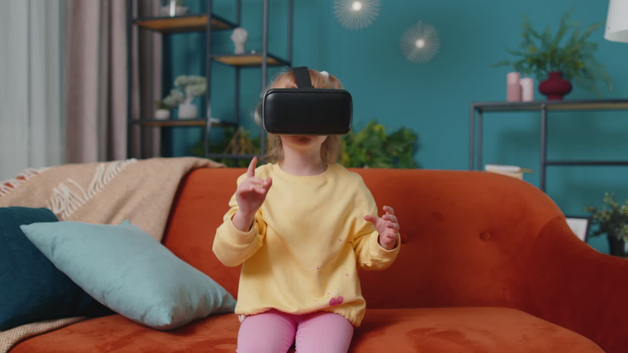 Toddler girl sitting on home sofa using virtual reality headset helmet app to play video 3D game