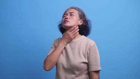asian middle age woman standing isolated on a blue background, hand to neck portraying painful sore throat symptom of flu infection.