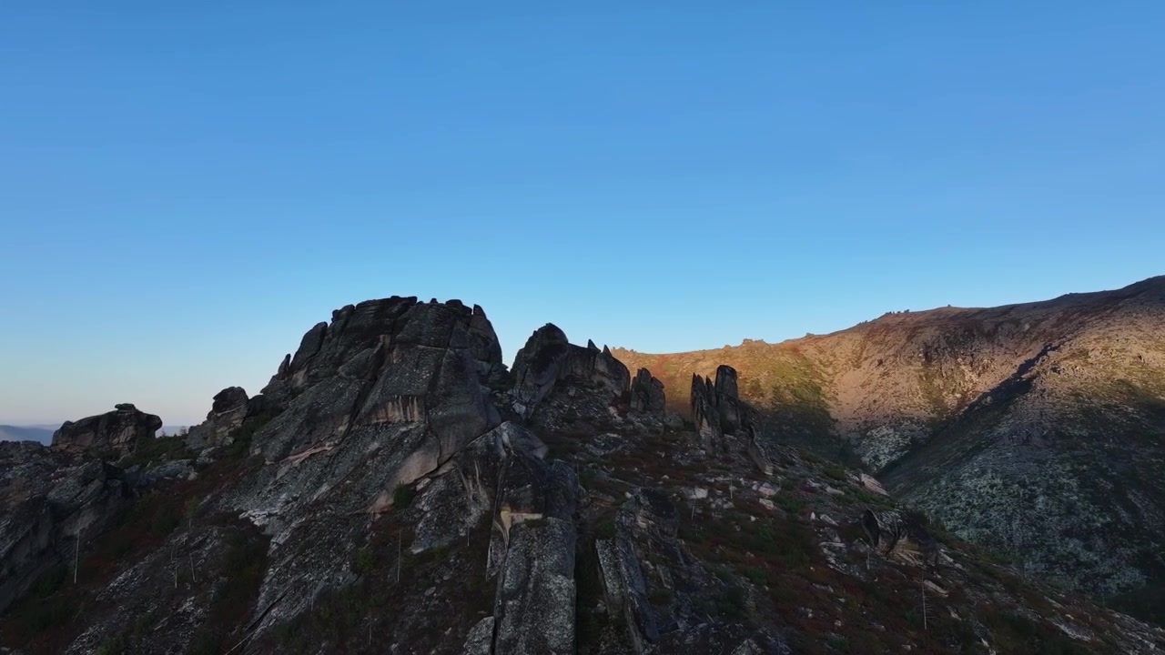 Veduta aerea di un aspro paesaggio montano con affioramenti rocciosi e piccoli edifici sotto un cielo azzurro limpido. La telecamera si muove lentamente per rivelare il fondovalle e le colline circostanti
