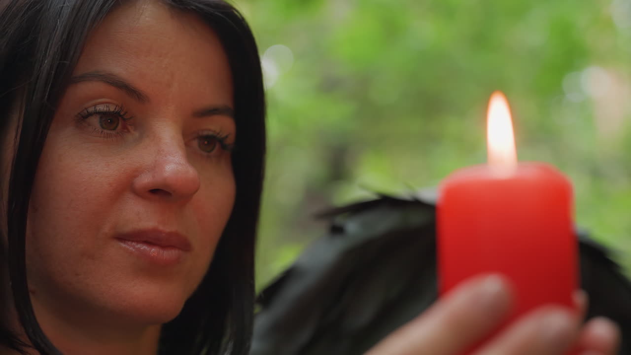 Primer plano de una vela roja encendida sostenida por una mujer bajo la suave luz de un bosque, con una llama cálida parpadeando sobre un fondo bokeh verde difuminado, evocando misterio, espiritualidad y conexión emocional con la naturaleza.
