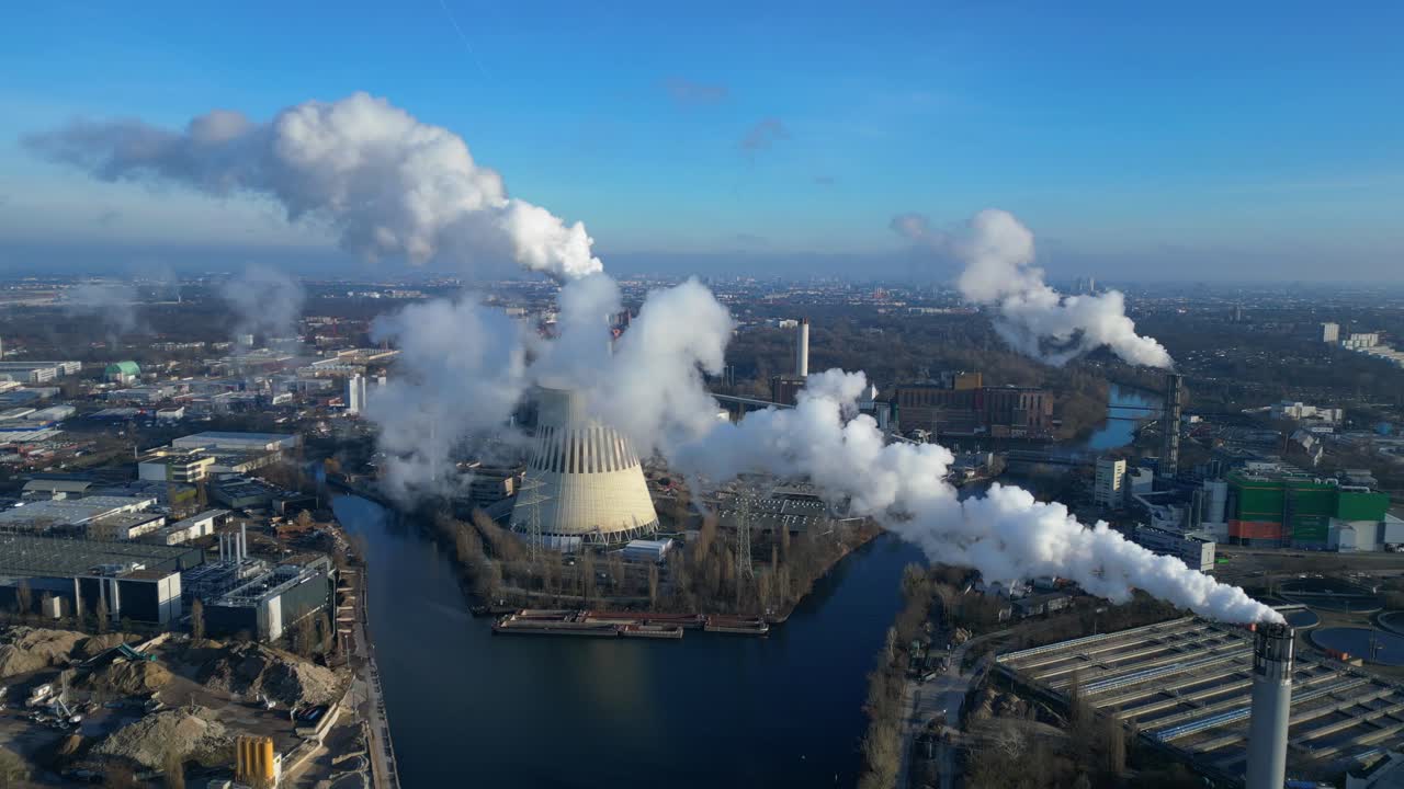 Aerial view of a thermal power plant emitting smoke on a sunny day, with a river and a city in the background. Amazing aerial view flight tilt up drone