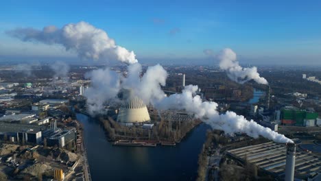aerial view of a thermal power plant emitting smoke on a sunny day, with a river and a city in the background. amazing aerial view flight tilt up drone