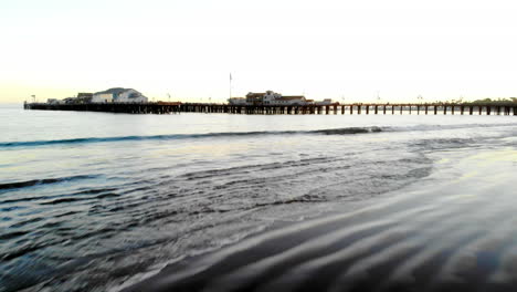 Luftdrohnenaufnahme,-Die-Tief-über-Die-Wellen-Und-Sandstrände-Fliegt-Und-Bei-Sonnenuntergang-In-Santa-Barbara,-Kalifornien,-Auf-Den-Stearns-Wharf-Pier-Voller-Touristen-Blickt