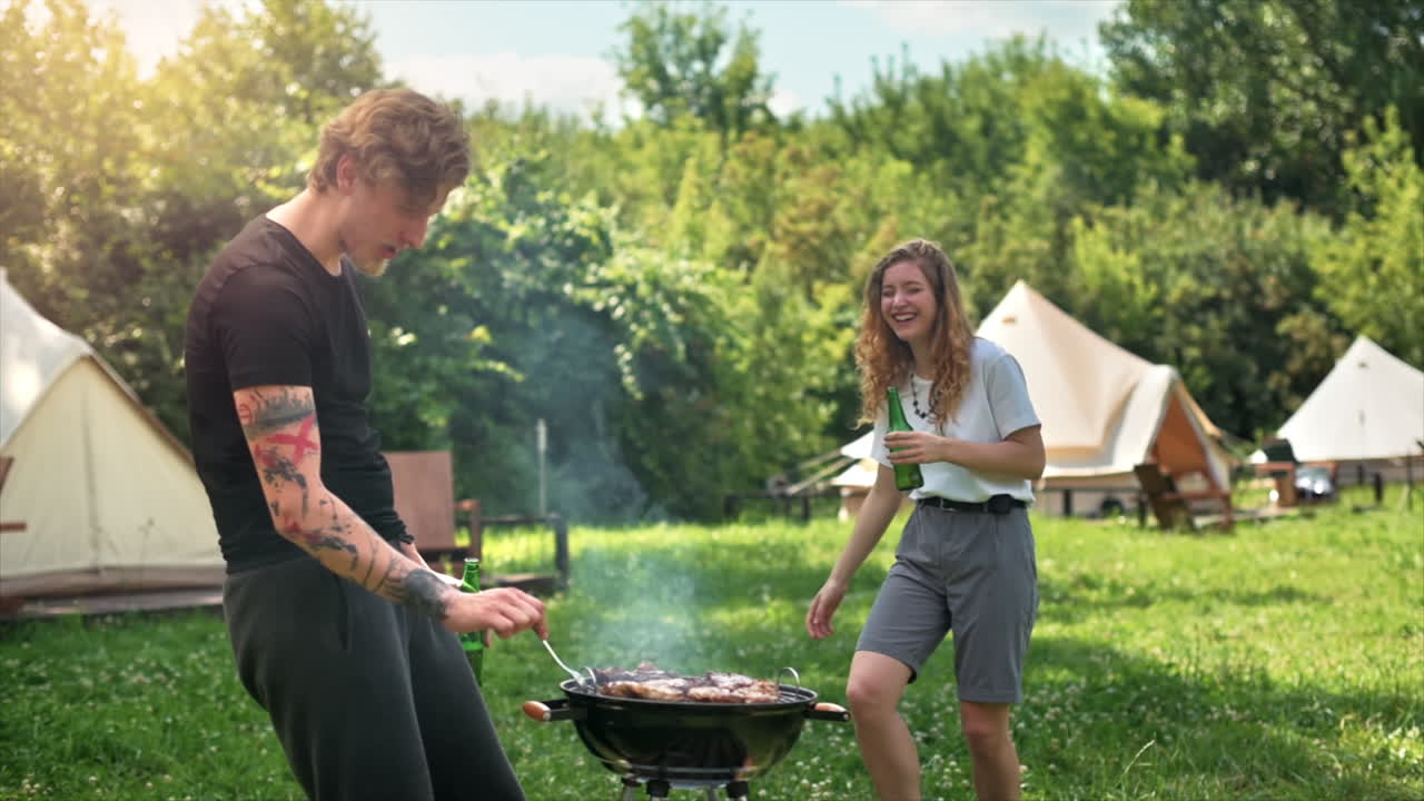 Young couple frying meat on the grill and drinking beer, laughing. Greenery around. Glamping. Slow motion