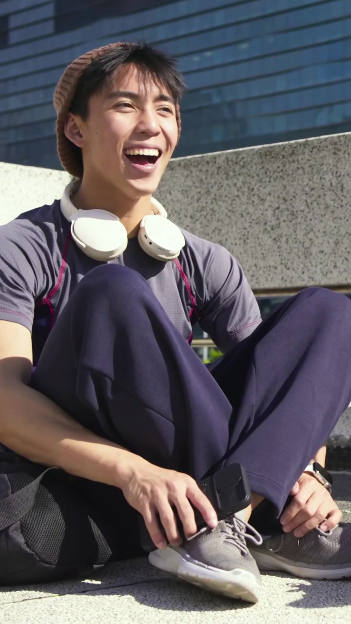 竖屏视频：亚洲男子戴着毛线帽和耳机，在台阶上查看手机，轻敲屏幕并展示给朋友看。