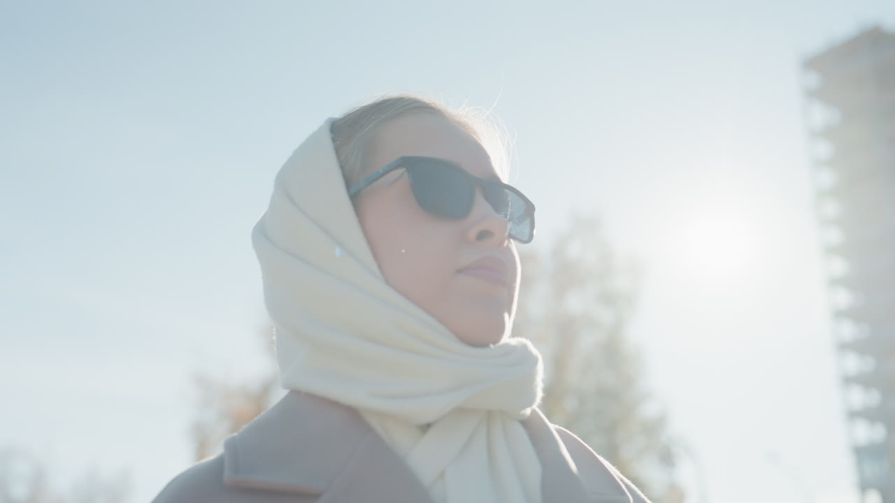 White woman wearing scarf sunglasses, vintage coat strolling autumn park under soft sunlight, adjusting frames, contemplative profile against city building backdrop, cozy wool texture, fashion