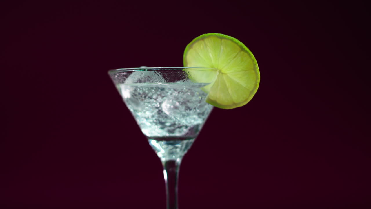 video del producto de un vaso de martini, gin tonic, bebida alcohólica con una rodaja de limón y bloques de hielo, girando en una mesa giratoria con fondo burdeos profundo