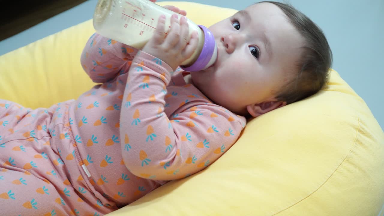 Bambina di dieci mesi che gioca con un biberon mentre beve latte artificiale.