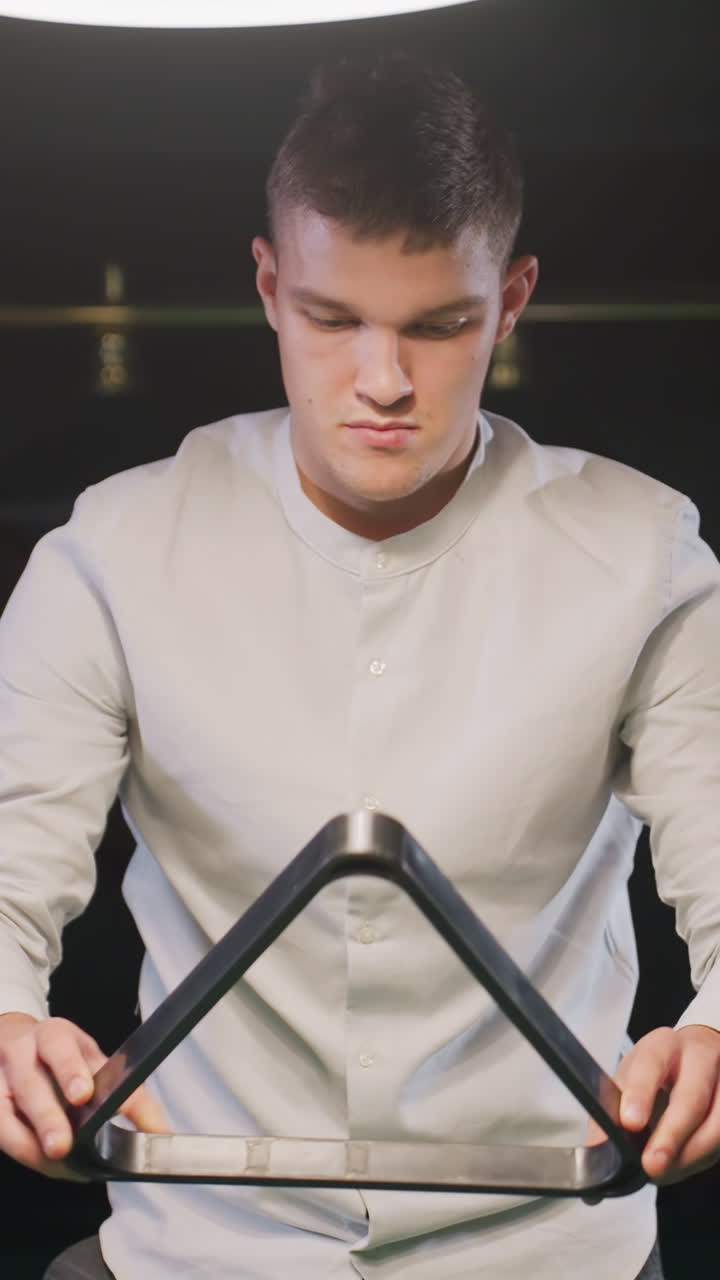 색 셔츠를 입은 젊은이가 따뜻한 환경 조명 아래 빌리어드 방에서 삼각형 랙을 들고 현대적인 장식으로 둘러싸인 정확하게 풀볼을 배열하는 데 집중하는 클로즈업