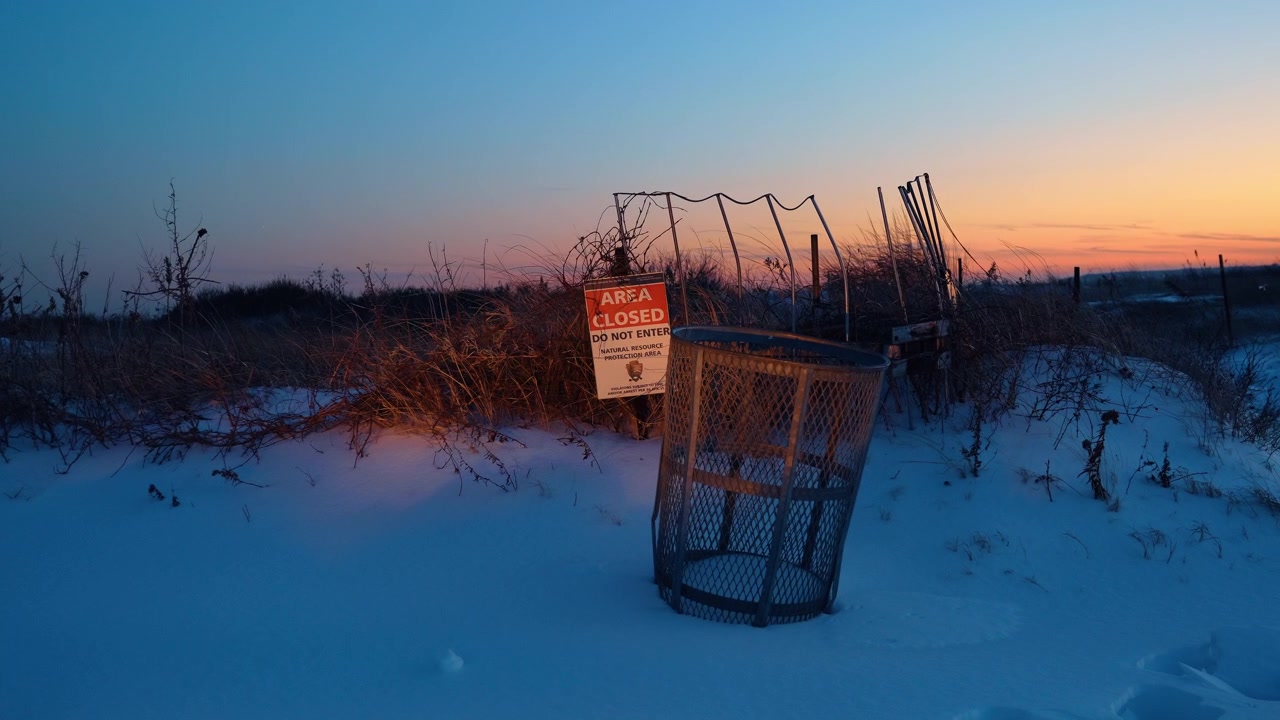 Area closed sign and trash bin in snow at sunset. A National Park Service restricted area sign and wire trash can on snowy dunes during winter sunset.