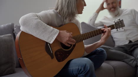 happy senior woman singing and playing the guitar sitting on chair, while in blurred background elderly friends listening to her and singing together sitting at the table 2