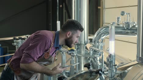 young male brewer wearing a leather apron supervise the process of beer fermentation at a modern brewery factory