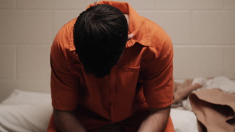 A-sad,-depressed-attractive,-handsome-man,-criminal-prisoner,-inmate-in-orange-jumpsuit-sitting-in-prison-cell-sitting-on-bed