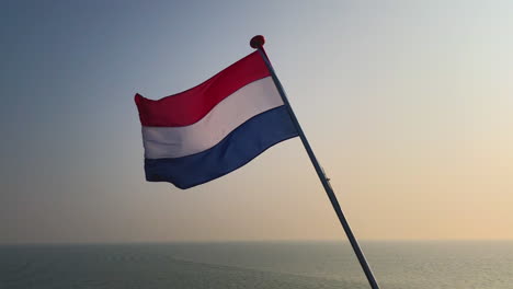 Niederländische-Flagge-Der-Niederlande-Im-Wind-Bei-Sonnenuntergang-Vor-Dem-Hintergrund-Fließenden-Wassers-Aus-Der-Nordsee