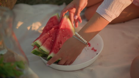 primer plano de la mano de una mujer que alcanza rebanadas frescas y jugosas de sandía en un plato blanco, la luz del sol proyecta sombras suaves, destacando los colores vibrantes de la fruta