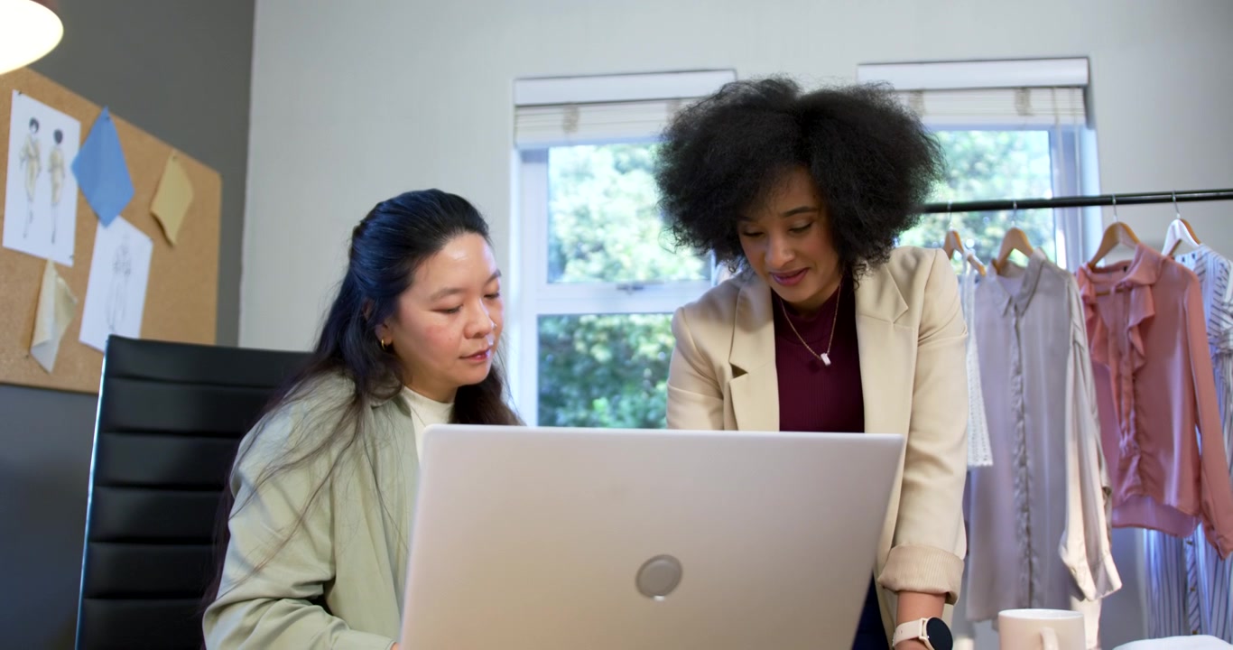 Diverse women leaning over laptop, reacting to mockup, reviewing sketches near clothing rack