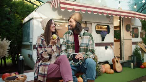 A-happy-brunette-girl-in-a-white-hat-holds-a-green-apple-in-her-hand-and-communicates-with-her-brunette-boyfriend-in-a-green-checkered-shirt-who-drinks-tea-and-sits-near-a-trailer-in-a-camp-outside-the-city-in-the-summer-during-a-picnic