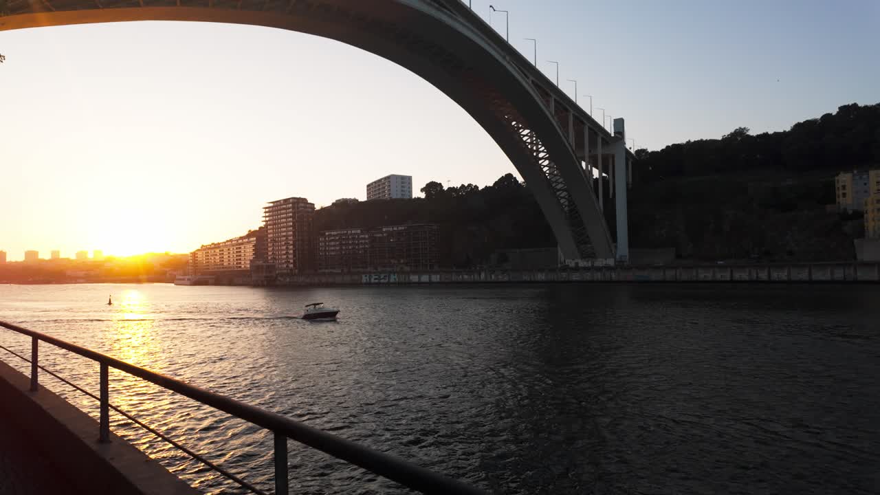 Sunset view of Douro River under a large bridge with buildings and a boat in Porto, Portugal