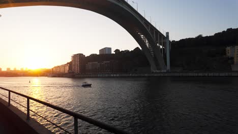 sunset view of douro river under a large bridge with buildings and a boat in porto, portugal