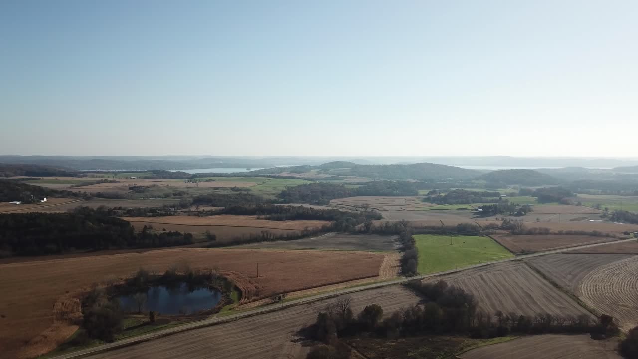 Aerial shot of rural farmland featuring a reflective pond, a red barn, patchworked fields, and rolling hills under a clear blue sky, depicting peaceful countryside living.