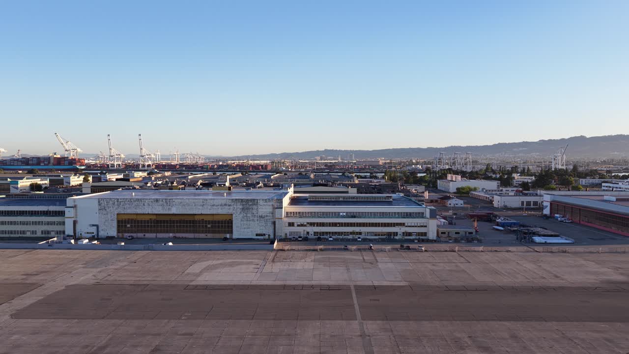 Vista aérea volando de lado sobre la antigua Estación Aeronaval de Alameda, mostrando los hangares históricos y las grúas del puerto de Oakland al otro lado del agua bajo un cielo azul claro al amanecer.