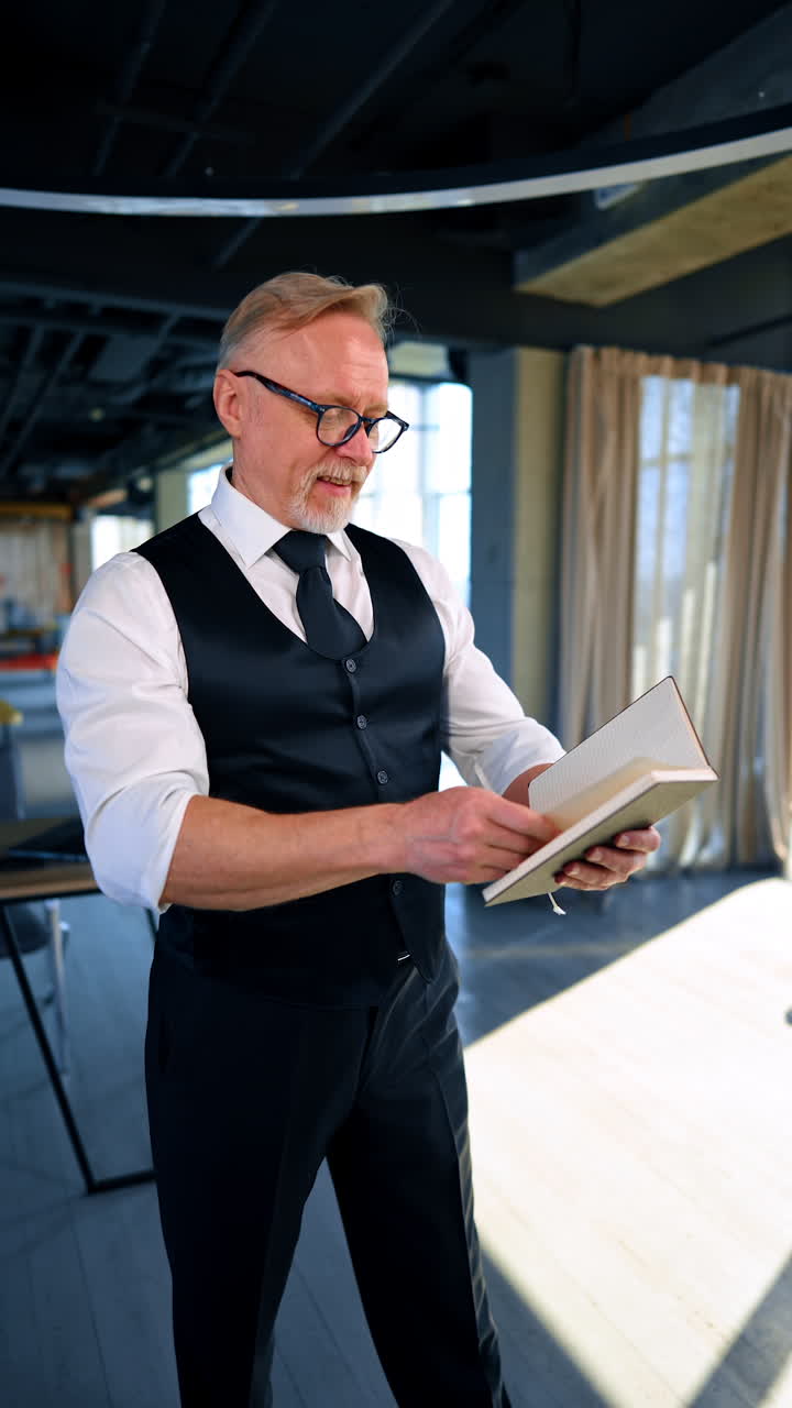 Positive mature man stands in the office. Man turns the pages of his notebook and then puts the book on his head. Vertical video.