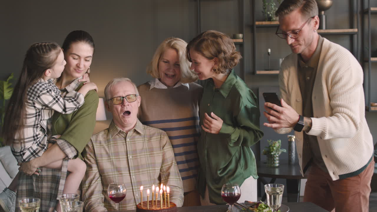 친척이 휴대전화로 비디오를 촬영하는 동안 집에서 가족과 함께 축하하는 동안 생일 케이크에 촛불을 끄는 노인