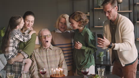 senior man blowing out candles on birthday cake during a celebration with his family at home while relative filming a video with mobile phone