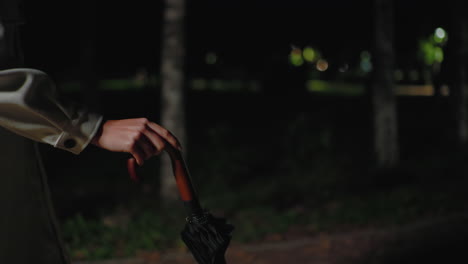 close up partial view of lady walking at night holding umbrella by wooden handle, wearing light coat, with blurry bokeh car lights in background and dim streetlight illuminating park pathway