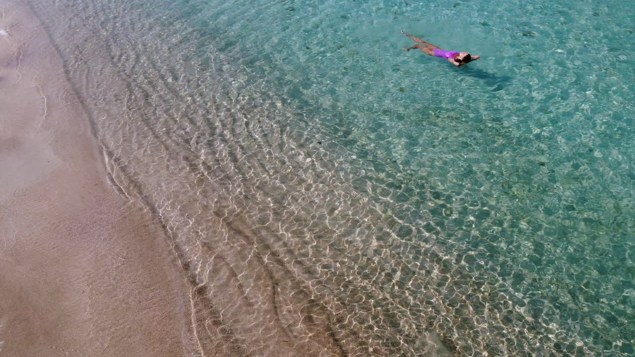 Aerial drone shot of Young woman dressed in pink swimsuit swimming in the crystal clear water of Diakofti beach, Kythira, Greece. Drone around the woman in the sea.