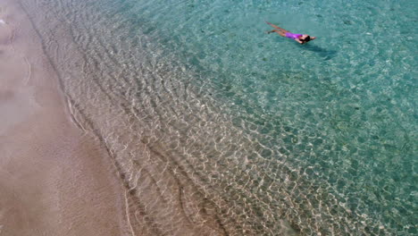 aerial drone shot of young woman dressed in pink swimsuit swimming in the crystal clear water of diakofti beach, kythira, greece. drone around the woman in the sea.