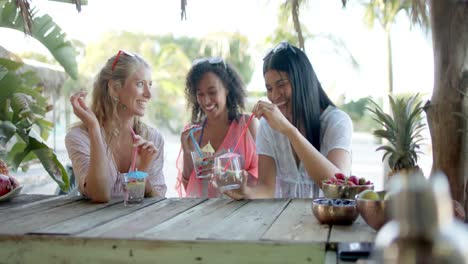 Happy-diverse-female-friends-drinking-cocktails-at-beach-bar-and-talking