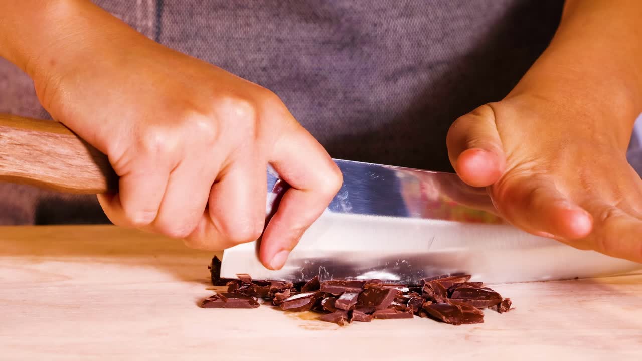 Close-up of hands skillfully chopping chocolate pieces with a large knife on a wooden cutting board.