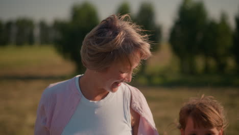 close-up of little girl walking alongside aunt as aunt smiles warmly and touches her hair in breezy farmland setting, background features blurred trees and open field under sunlight