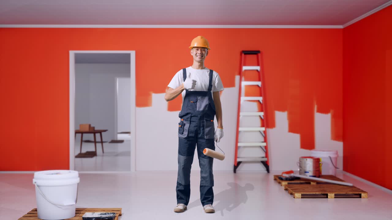 cuerpo lleno de pintor hombre asiático con casco de seguridad sonriendo y mostrando pulgares hacia arriba gesto mientras estaba de pie con la pared de pintura en la habitación