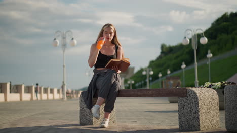 woman seated outdoors holding book in one hand while sipping juice from bottle in the other, continuing to read, blurred background includes lamp posts, greenery, and people walking