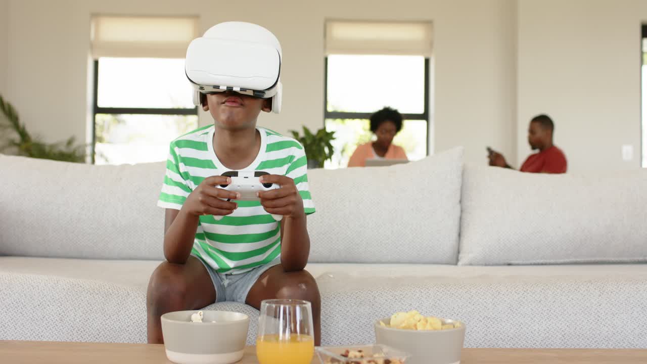 African American boy playing VR game holding controller on sofa while parents scrolling smartphones
