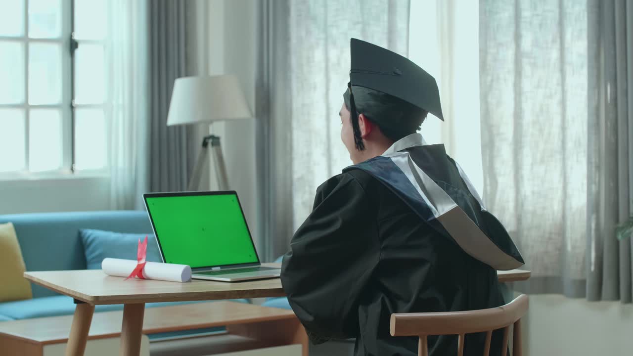 Excited Asian Man Having Video Call On Laptop Computer With Green Mock-Up Screen. Male Graduate Wearing A Graduation Gown And Cap Sitting On The Living Room