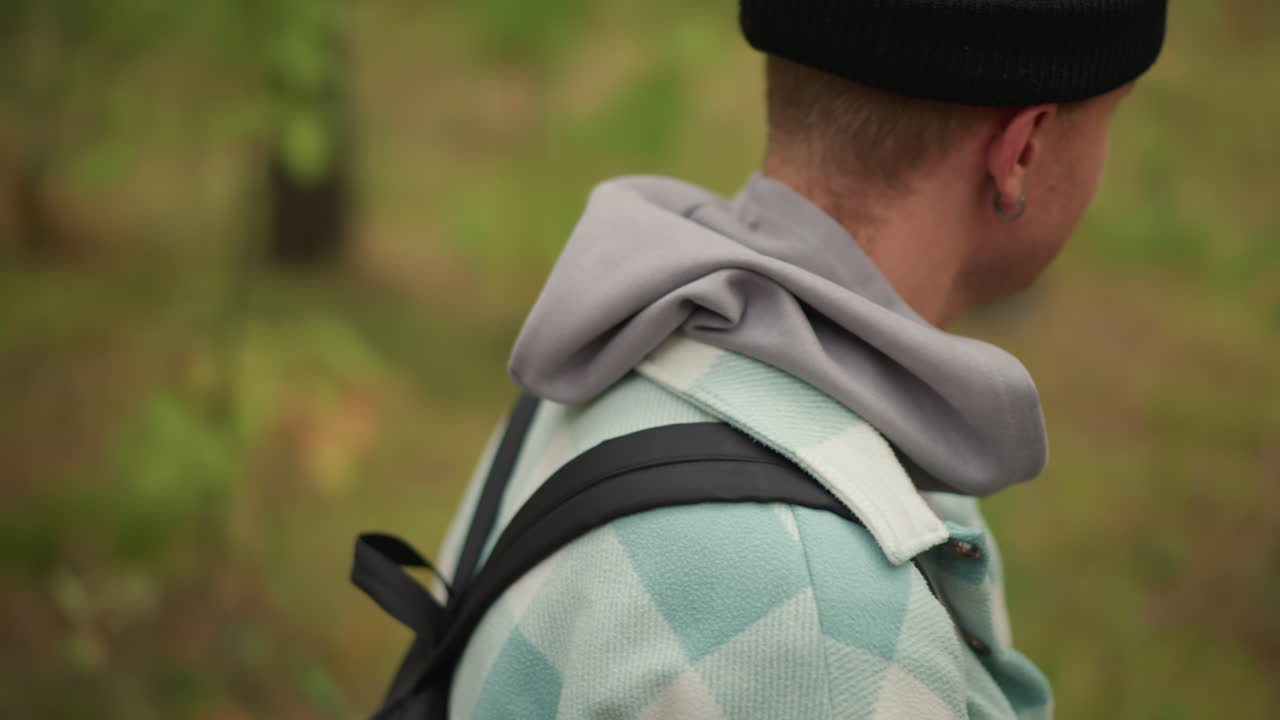 Vit backpacker utforskar en stilla skogsstig, ser sig omkring på trädstammar och undervegetation, huvtröja och rutig jacka, stadigt tempo och nyfiken blick, öppen naturdokumentärkänsla