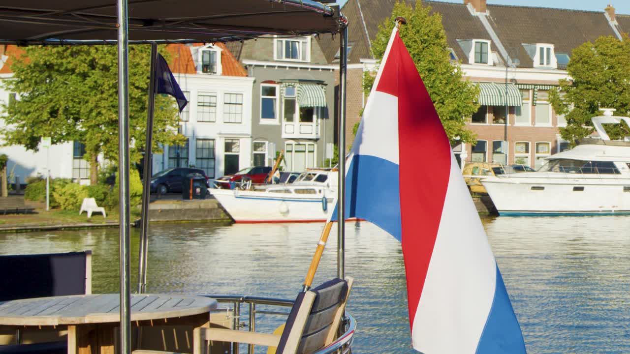 A Dutch flag flutters in the breeze beside a canal in Haarlem, with historic buildings, boats, and bright daylight creating a lively urban scene